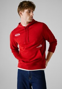 Roter Kapuzenpullover mit einer Fronttasche, verstellbarer Kapuze und weißen Logodetails. Kombiniert mit dunkelblauen Jeans, die über eine weiche Textur und ein lässiges Design verfügen.