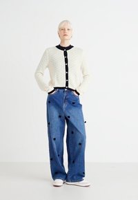 Cardigan crem cu nasturi negri și detalii negre, asortat cu blugi din denim albastru cu picioare largi, având accente cu pompoane negre. Incaltaminte alba.