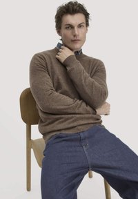 Brauner Wollpullover mit Rundhalsausschnitt, über einem karierten Hemd getragen, kombiniert mit blauen Denim-Jeans. Das Model sitzt auf einem Holzstuhl, die Hand in der Nähe des Nackens.