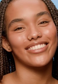 Portrait d'une personne avec des cheveux tressés, portant une boucle d'oreille en or et de petites boucles d'oreilles triangulaires, souriant avec une peau lisse et claire.