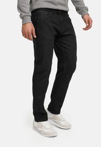 Mann iført svarte jeans, grå langermet skjorte og beige og hvite joggesko, stående med armene avslappet langs siden.