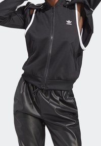 Schwarze Kapuzenjacke mit weißen Akzenten und Logo, mit Kapuze und offenen Ärmeln, kombiniert mit glänzenden schwarzen Hosen. Glatte Stoffstruktur.