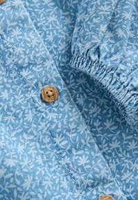 Tissu à motif floral bleu avec de petites feuilles blanches, présentant un bouton en bois et un détail de manche froncée. Matériau texturé et léger.