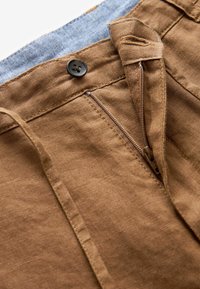 Pantalon en lin brun avec une taille en denim bleu, présentant une fermeture éclair à l'avant et un bouton, des passants de ceinture sur les côtés et un tissu texturé.