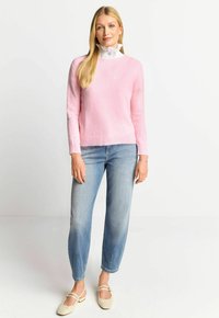 Rosa gestrickter Pullover mit Rundhalsausschnitt, kombiniert mit hellblauen geraden Jeans und beigen Riemchenschuhen. Ein weißer Rüschensaum ist sichtbar.