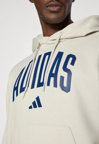 Bežna kapuca iz mešanice bombaža, z velikim modrim logotipom "ADIDAS" v krepkem fontu in majhnim logotipom trefoil Adidas na sprednji strani.