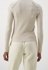 Person som bär en beige, långärmad ribbad topp med perforerade detaljer på ryggen och krämfärgade byxor, stående mot en enfärgad bakgrund.