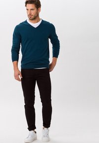 Maglione teal a scollo a V, con polsini e orlo a costine, abbinato a pantaloni neri slim-fit e sneakers bianche, outfit casual su un modello.