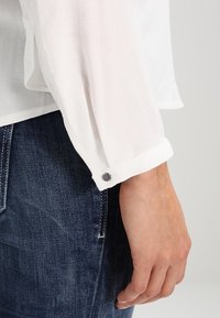 Vit, genomskinlig långärmad skjorta med uppvikta manschetter och dekorativ knappdetalj. Bärs med mörka denimjeans som visar mjuk tygstruktur.