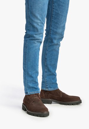 Bottines en suède marron avec une finition texturée et des semelles en caoutchouc, associées à un jean en denim bleu à coupe slim.