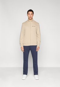 Sweat-shirt beige zippé avec col montant, arborant un logo brodé noir, associé à un pantalon bleu marine et des baskets blanches. Texture de tissu lisse.