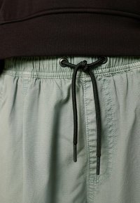 Hellgrüne Shorts mit einer strukturierten Oberfläche, einem elastischen Bund mit schwarzem Kordelzug und einem oben sichtbaren schwarzen Oberteil.