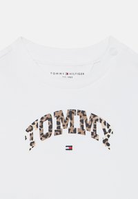 Balts kokvilnas T-krekls ar leopardu rakstu "TOMMY" logotipu, ar mazu sarkanu, baltu un zilu akcentu zem teksta un spiedpogām uz pleca.