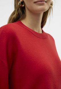 Pull rouge tricoté avec un col rond et des épaules tombantes. Présente une finition texturée et est assorti à de grandes boucles d'oreilles dorées.