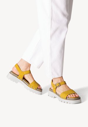 Gelbe Wildledersandalen mit verstellbarem Knöchelriemen, Gummisohle und Metallnietenakzenten. Sie verfügen über ein offenes Toe-Design und einen stabileren Absatz.