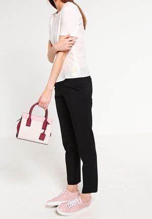 Femme portant un haut blanc à manches courtes, un pantalon noir et des baskets roses, tenant un sac à main bicolore rose par ses poignées sur fond blanc.