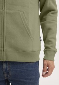 Felpa verde realizzata in morbido tessuto, con chiusura a zip, tasche a marsupio e polsini a coste. Abbinata a jeans in denim blu.