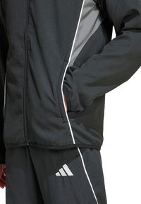 Schwarze Zip-Jacke mit grauen Akzenten und weißen Paspeln, gefertigt aus einem strukturierten Stoff und mit Seitentaschen. Kombiniert mit passenden Hosen.