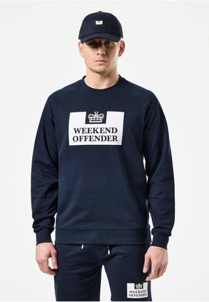Muž nosiaci tmavomodrú šiltovku, mikinu a nohavice, všetko s bielym logom "Weekend Offender" a grafikou koruny, stojaci na bielom pozadí.