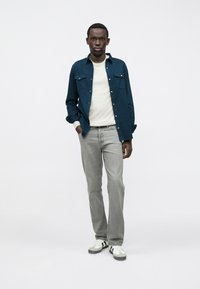 Man staat met zijn handen in zijn zakken, draagt een witte trui, een donkerblauw overhemd met knopen, grijze spijkerbroek en witte sneakers met zwarte strepen.