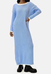 Robe longue tricotée côtelée bleu clair avec un large décolleté, des manches longues, et un liseré blanc au niveau du col et des poignets, portée avec des bottes à plateforme noires.