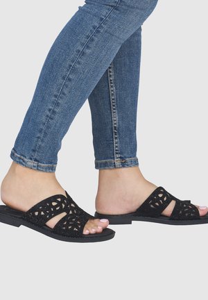 Pieds portant des sandales noires ouvertes à enfiler avec des motifs ajourés, associés à un jean skinny en denim bleu sur un fond uni.