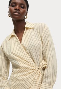 Camicia a righe gialla chiaro, con colletto, vita annodata e maniche lunghe, caratterizzata da sottili righe verticali nere.