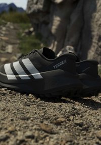 Черно-бели маратонки Adidas Terrex за бягане по пътеки на скалиста земя с размит скалист терен на заден план.