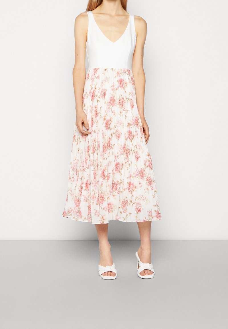Robe florale avec un buste ajusté blanc et une jupe plissée, longueur cheville, ornée de fleurs roses sur un fond clair, associée à des sandales blanches.
