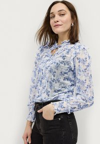 Leichte, durchsichtige Bluse mit blauem Blumenmuster, Rüschensaum an Kragen und Ärmeln, Schlüssellochdetail am Ausschnitt, kombiniert mit dunklen Jeans.