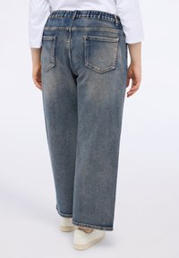Pantalones de mezclilla de pierna ancha en azul claro con una cinturilla elástica, dos bolsillos traseros y dobladillo sin terminar. Combinados con zapatillas blancas.