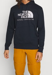 Osoba v černé mikině The North Face s logem a hořčicově žlutých kalhotách, postávající před jednoduchým světlým pozadím.