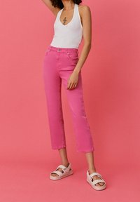 Pantalon en denim rose vif taille haute avec jambe droite, deux poches avant et un seul bouton sur le dessus. Porté avec des sandales blanches.