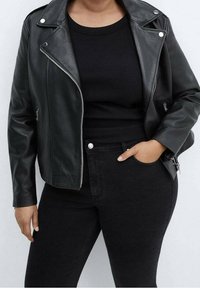 Chaqueta de cuero negra de motorista con herrajes plateados, bolsillos con cremallera y una silueta ajustada. Llevada sobre una camiseta negra con relieve y vaqueros.