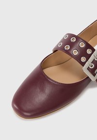 Chaussure en cuir bordeaux avec un bout carré, dotée d'une large sangle avec des découpes circulaires et des détails en boucle en argent. Semelle plate.