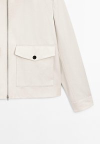 Lys beige jakke med lynlås, en enkelt brystlomme med en sort knap og en afslappet pasform med synlige stikninger.