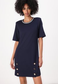 Navy kortermet kjole med rund hals, med beige detaljer og dekorative knapper langs den ene siden. Myk, glatt stofftekstur.