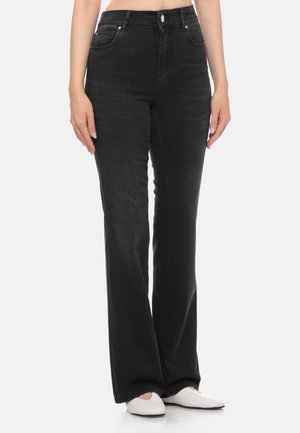 Donna con jeans neri dritti a vita alta e ballerine bianche a punta, in piedi su sfondo semplice.