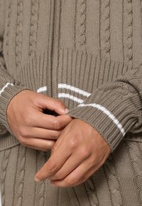 Barna, kötött pulóver, amely kábelmintás textúrával és fehér csíkos akcentussal rendelkezik a mandzsettákon, ribbelt szegéllyel és testhez simuló ujjal.