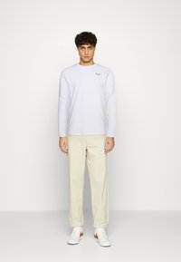 Langärmliges weißes T-Shirt mit einem leichten Stretch und kleinem schwarzem Logo, kombiniert mit hellbeigen Hosen und weißen Sneakers mit orangenen Akzenten.