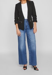 Veste noire avec poches zippées, associée à un jean bleu à jambes larges. Le look comprend des talons noirs ouverts à l'avant, mettant en valeur un style décontracté.