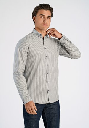 Mand iført en grå og hvid vertikalstribet skjorte med knapper og mørkeblå jeans, der justerer skjortekraven med højre hånd.