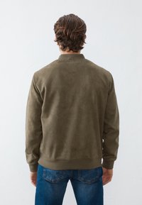 Oliwkowa zamszowa kurtka bomberka z prążkowanym kołnierzem i mankietami, widok z tyłu, w zestawieniu z niebieskimi dżinsami. Gładka tekstura, minimalistyczny design.