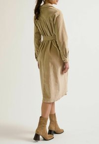 Eine Frau steht mit dem Rücken zum Betrachter, trägt ein langes beiges Kleid und braune Knöchelstiefel, vor einem neutralen Hintergrund.