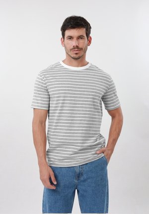 Homme aux cheveux courts et foncés portant un T-shirt à rayures grises et blanches et un jean bleu, debout avec une main dans sa poche.