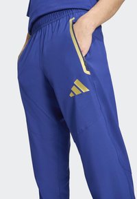 Niebieskie spodnie sportowe wykonane z gładkiej tkaniny, z suwakiem w kieszeni i złotymi detalami, w tym logo Adidas na dolnej części nogawki.