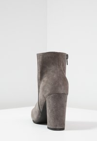 Bottine en daim gris avec un talon carré haut, présentant une texture lisse et une fermeture éclair latérale pour un accès facile. Se termine par un bout arrondi.