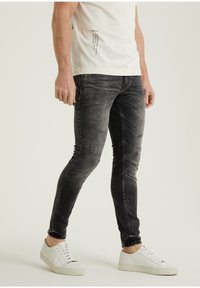 Mies, joka pukeutunut haalistuneisiin mustiin skinny-farkkuihin, valkoiseen painettuun t-paitaan ja valkoisiin rentoihin lenkkareihin, seisoo yksiväristä taustaa vasten.