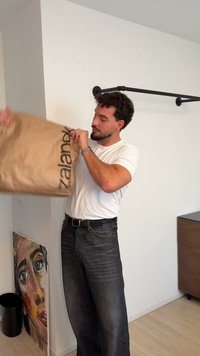 Smeđa papirnata vrećica za kupovinu s natpisom „Zalando“ u crnoj boji, koju drži osoba odjevena u bijelu majicu i tamne traperice u minimalističkoj sobi.