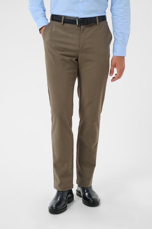 Hombre con pantalones marrones, cinturón negro, camisa azul claro y zapatos de vestir negros, de pie con una mano en el bolsillo.